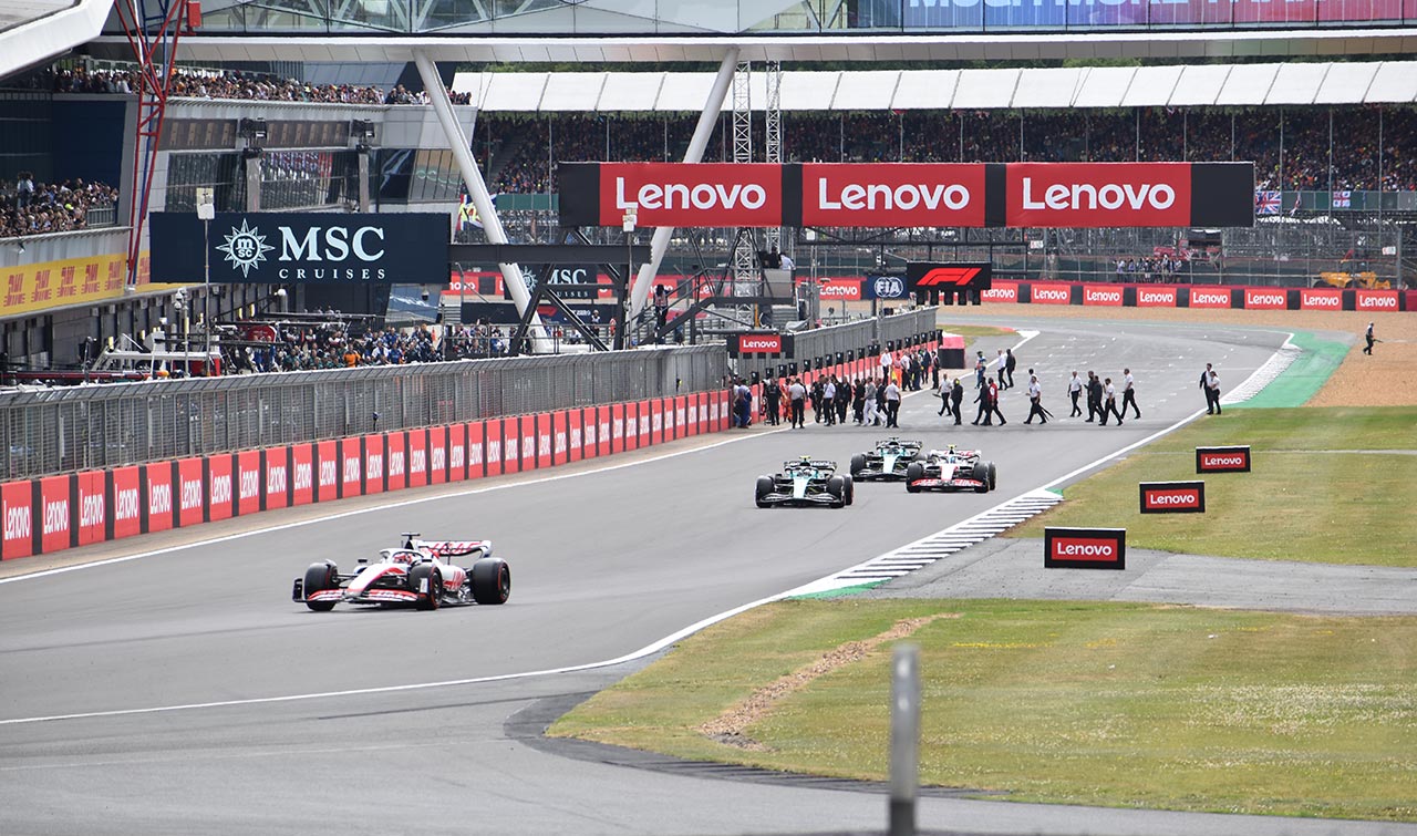 4 racing cars at the first turn with stadium and pits in the background