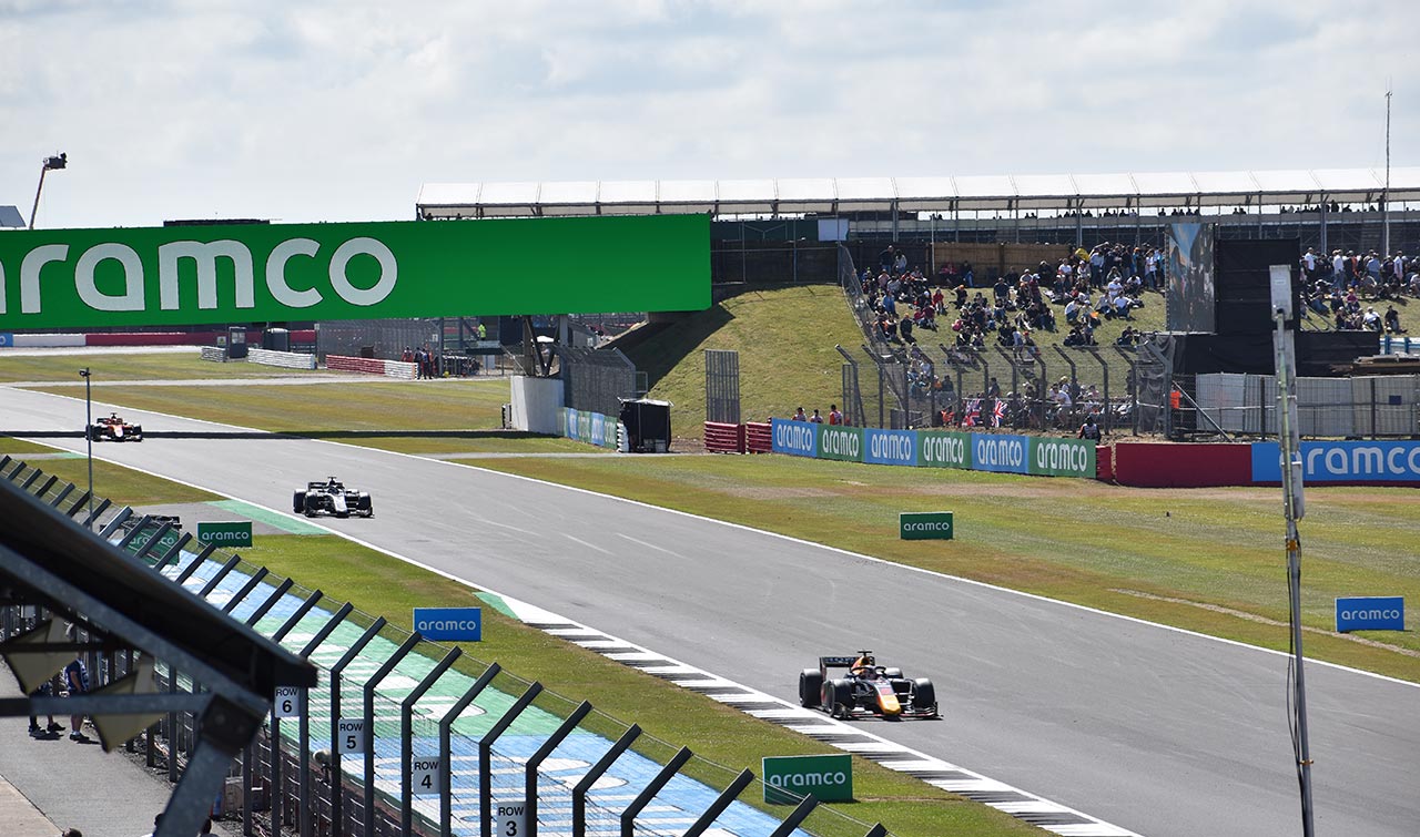3 f1 cars racing with grass seating with crowd in the background