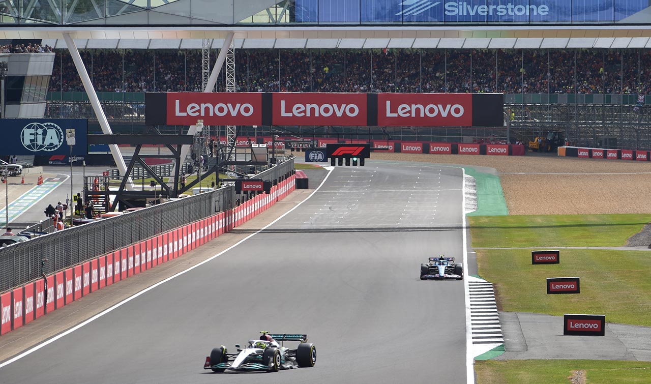 2 f1 cars racing by the startline with a big silverstone sign and crowds in the picture