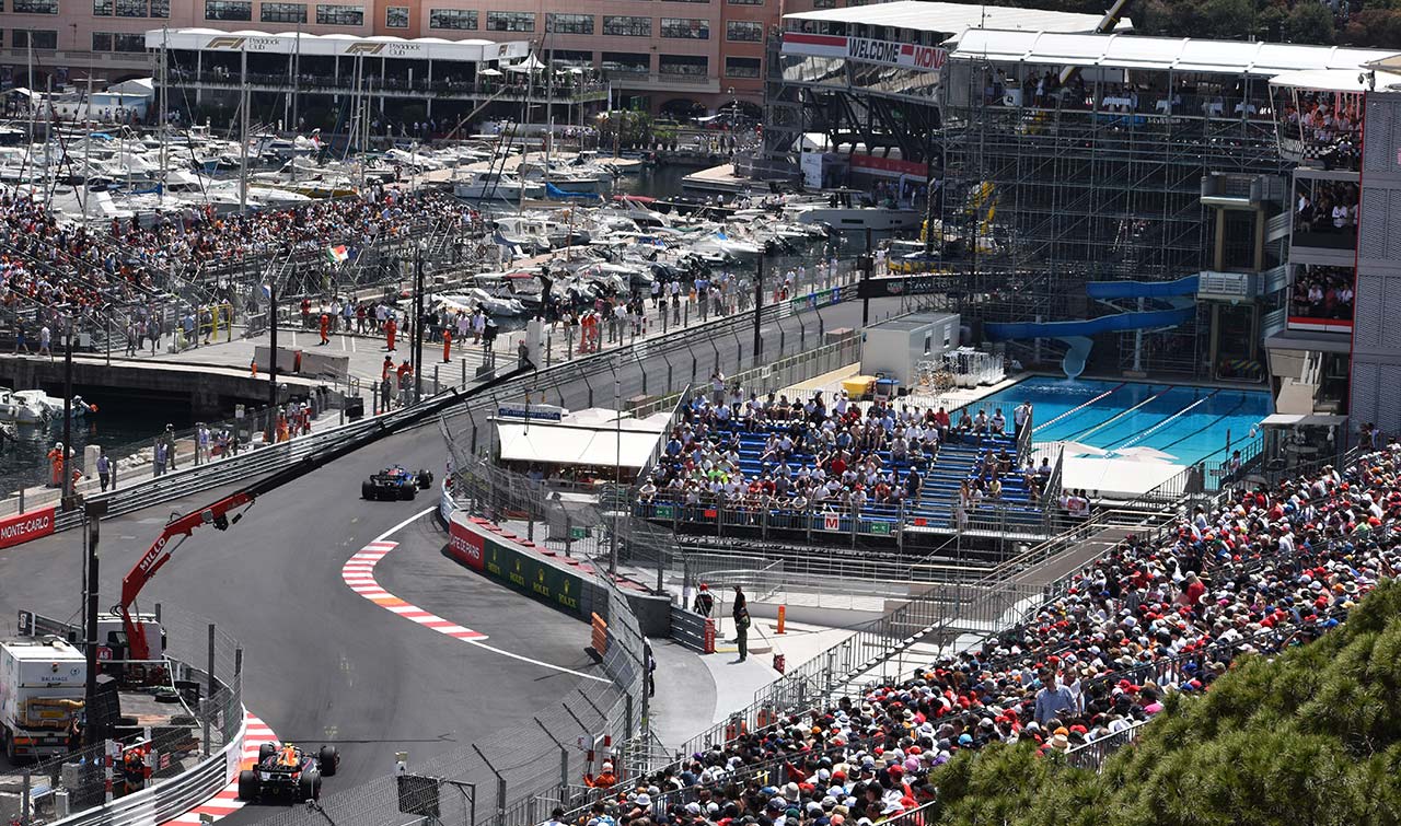 2 F1 cars racing at the monaco gp with the famous swimming pool in view and fans in grandstands all around