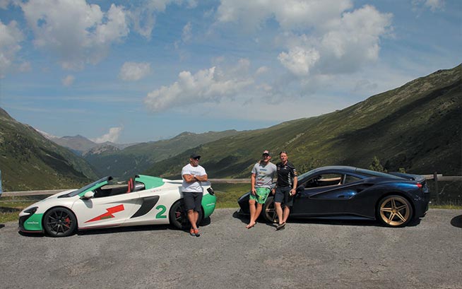 friends standing beside 2 sports cars with a valley in the background