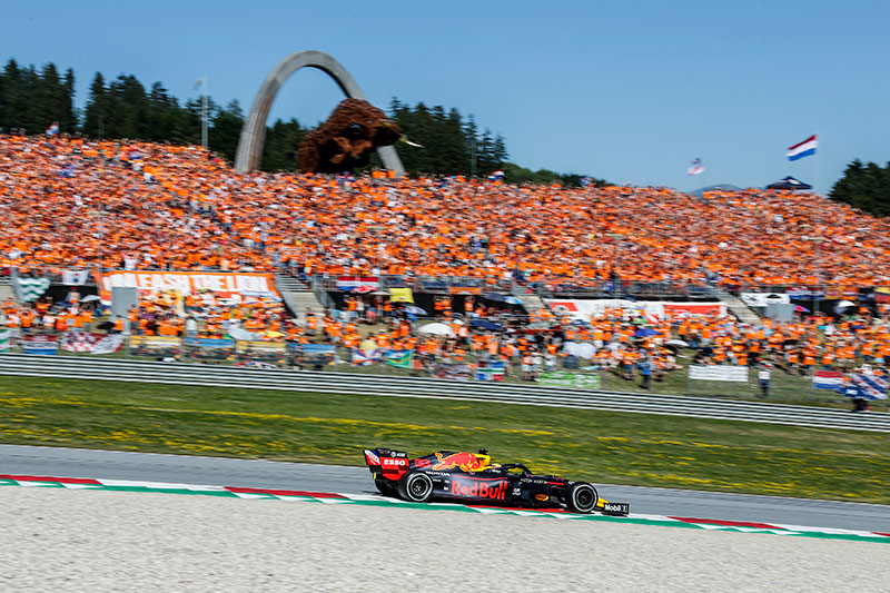 large crowd in orange at the austrian grand prix with a metal bull in the background and a f1 car in the foreground