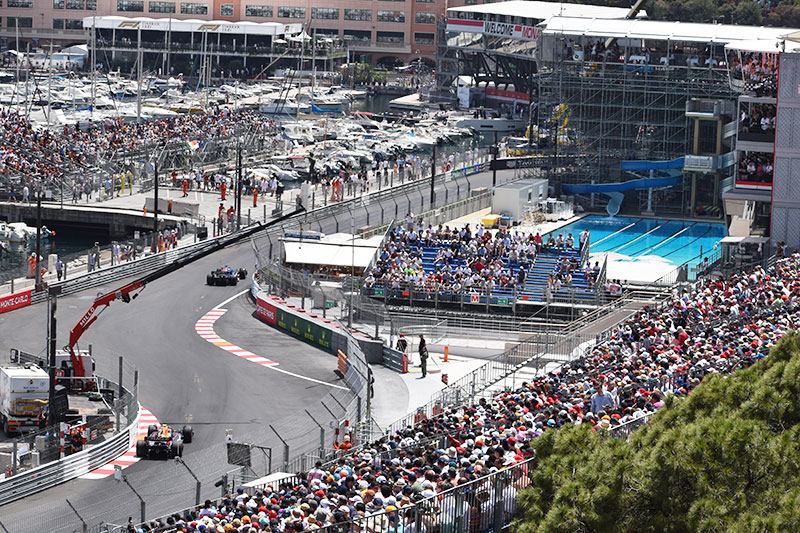 2 f1 cars racing in monaco with the crowds watching and view of the famous swimming pool